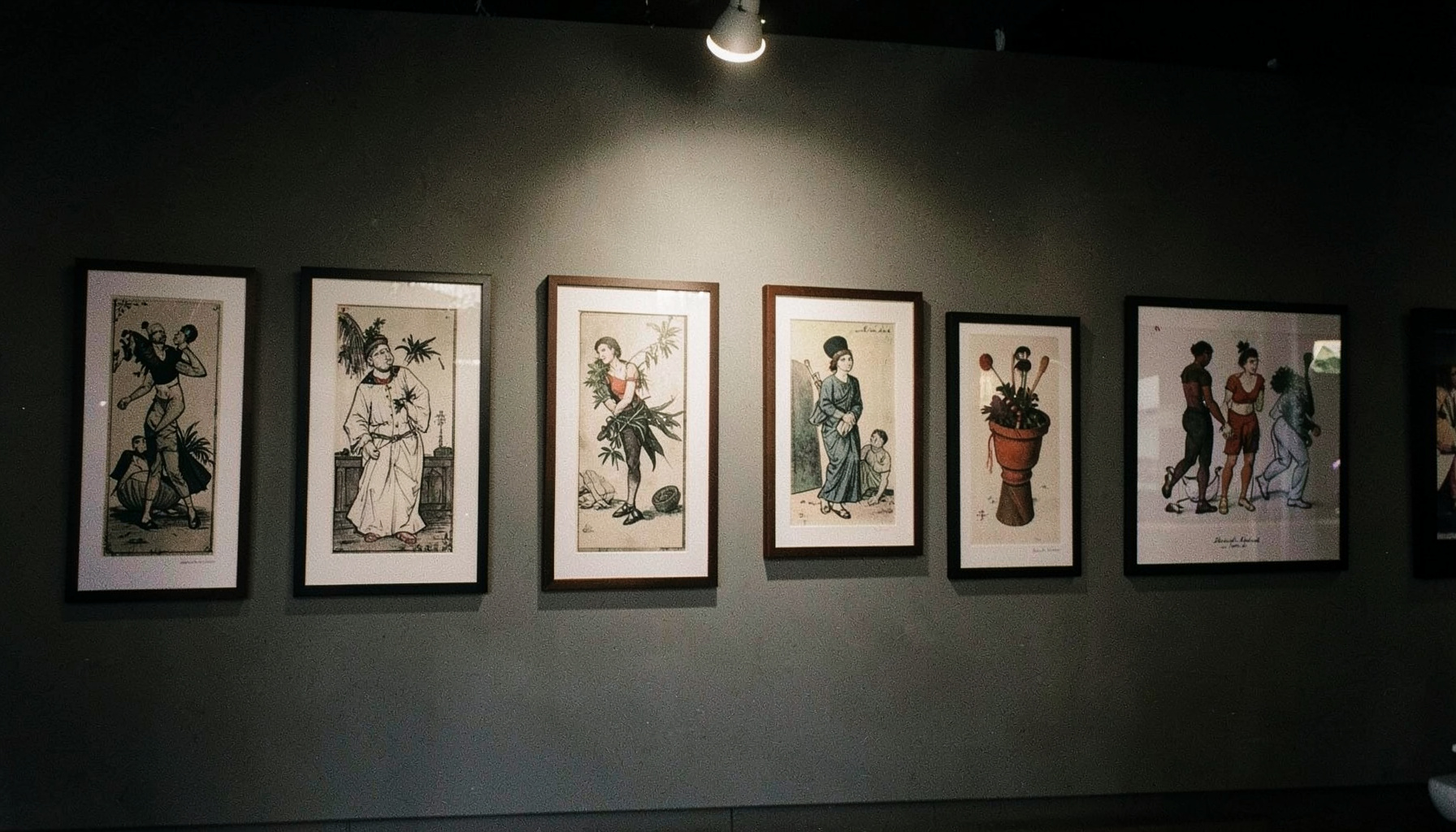 Gallery wall with framed illustrations lit by a single soft overhead light, dark and gallery-like mood