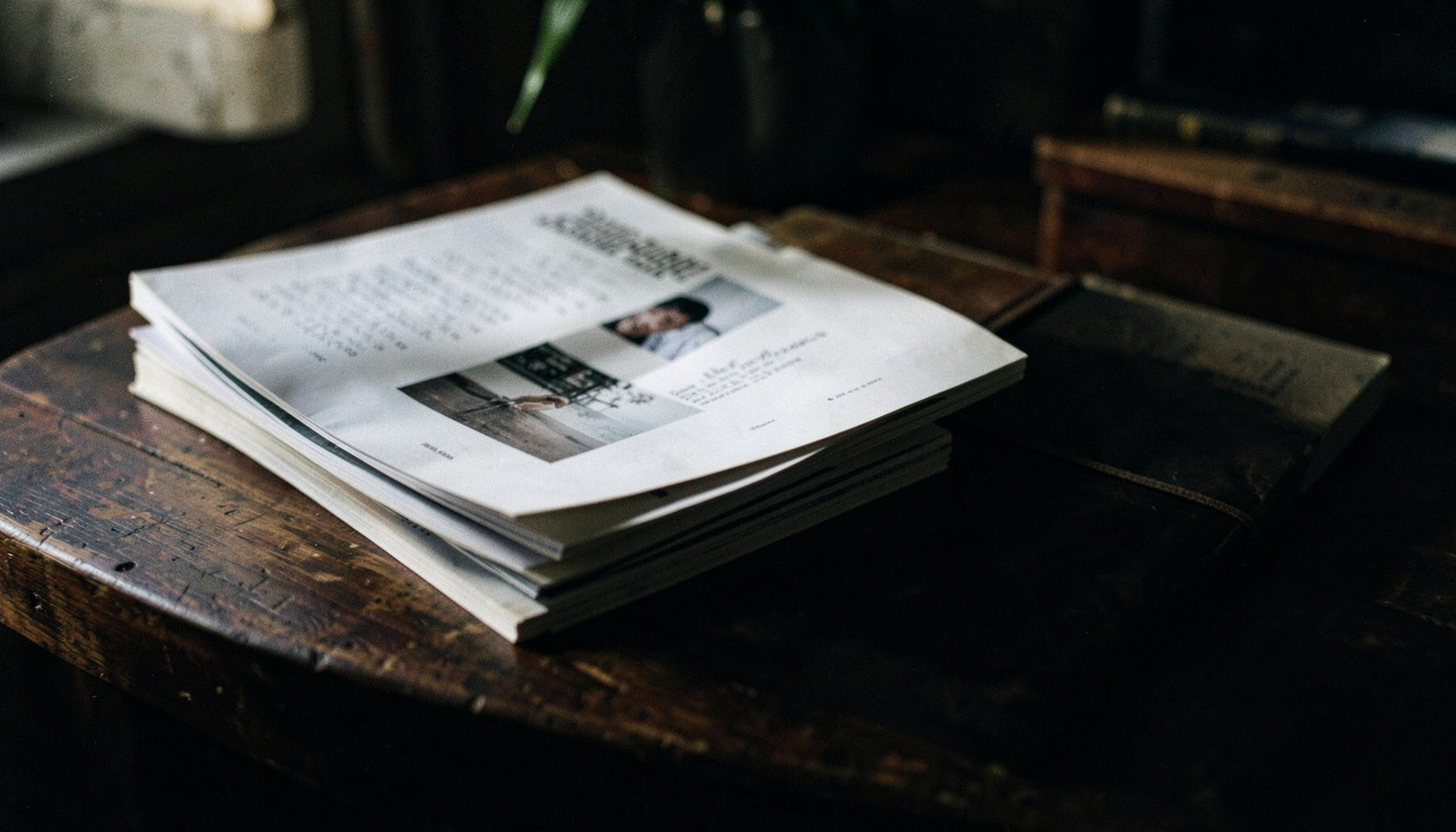 Stack of printed magazines and bound issues on a dark wooden table, moody low light, literary atmosphere
